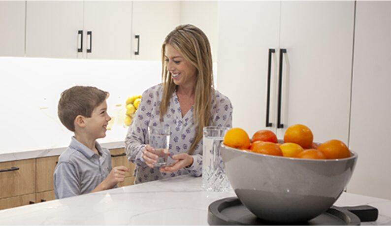 Mom pouring water for her son at the kitchen counter beside a bowl of oranges